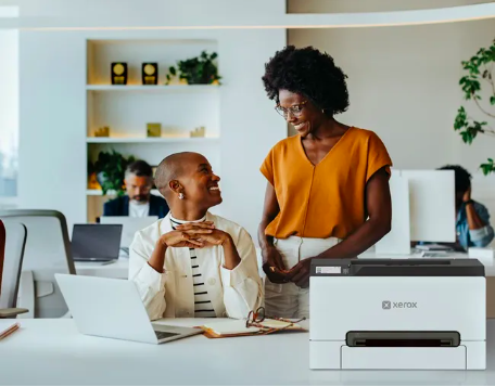 Deux femmes sourient en open space, sur le bureau une Imprimante couleur Xerox® C240.
